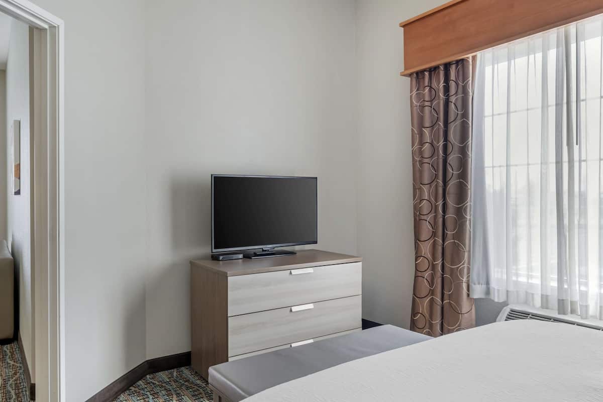 A hotel room with a bed, a TV on a wooden dresser, a window with curtains, and a door.