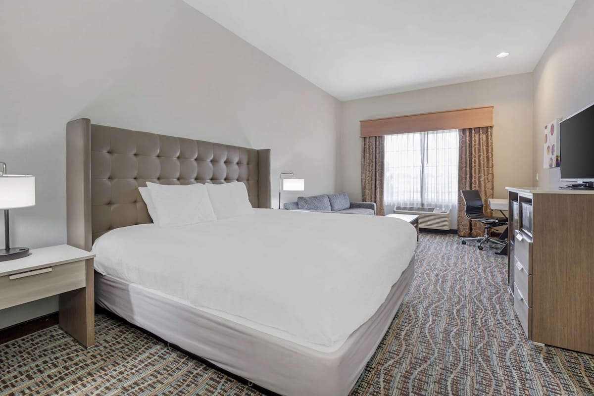 A hotel room with a large bed, a desk with a lamp, a TV, and a seating area with a chair and a sofa.