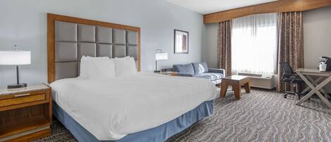 A hotel room with a large bed, a desk, a chair, a sofa, and a window with curtains.