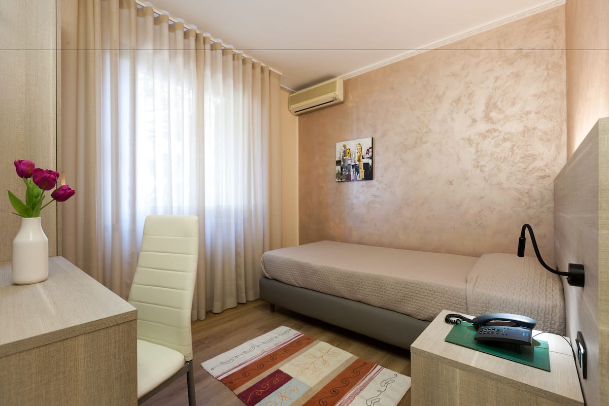 Een kleine, verzorgde kamer met een bed, een bureau met een bloevas, een stoel en een aan de muur bevestigde telefoon.