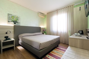 Habitación de hotel ordenada con cama, mesita de noche, escritorio y una planta pequeña.