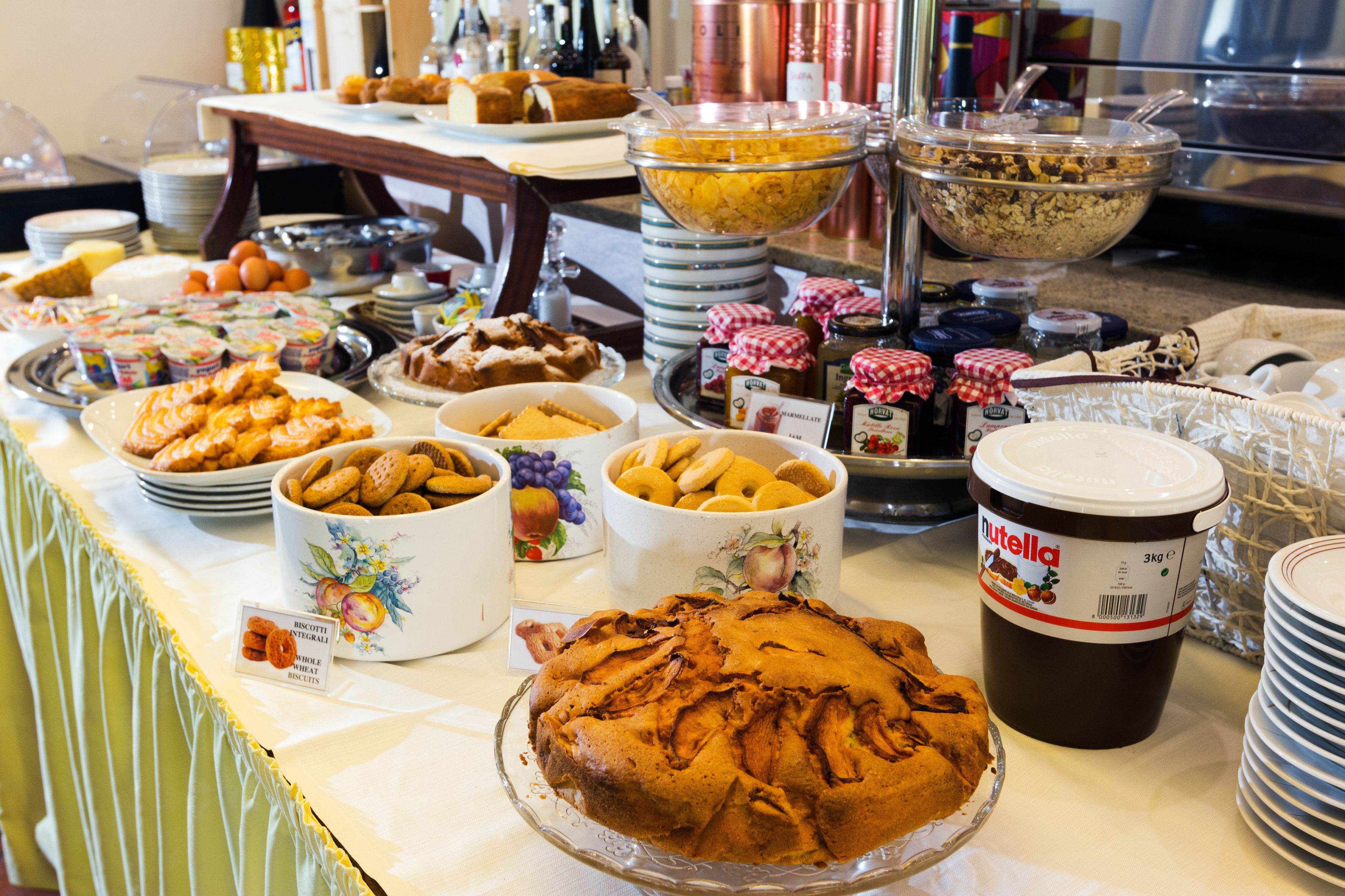 Una mesa de buffet con diversos alimentos, incluyendo un pastel grande, tazones de frutos secos y un tarro de Nutella.
