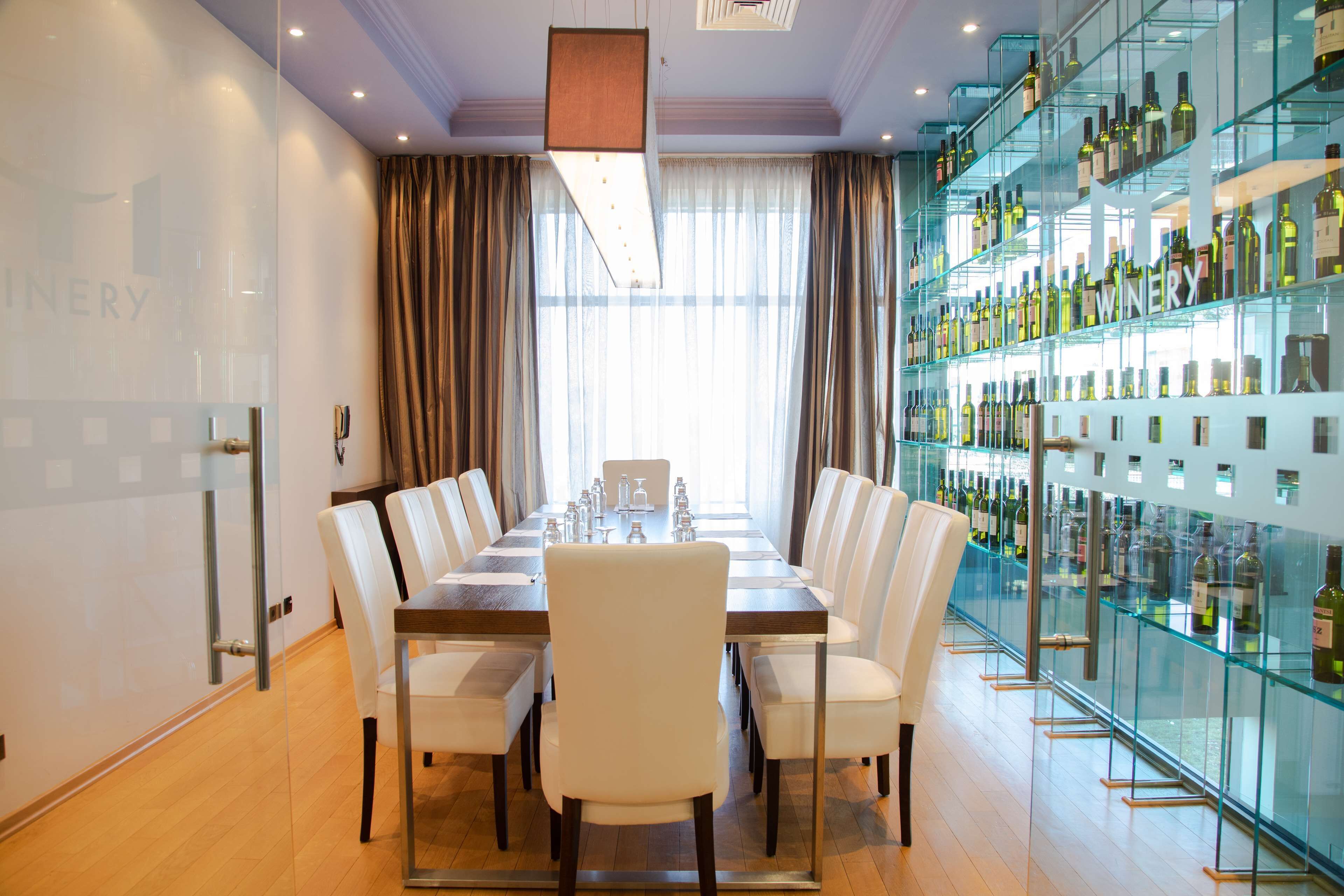 A modern dining area with a long table, white chairs, and a glass wine display on the right.