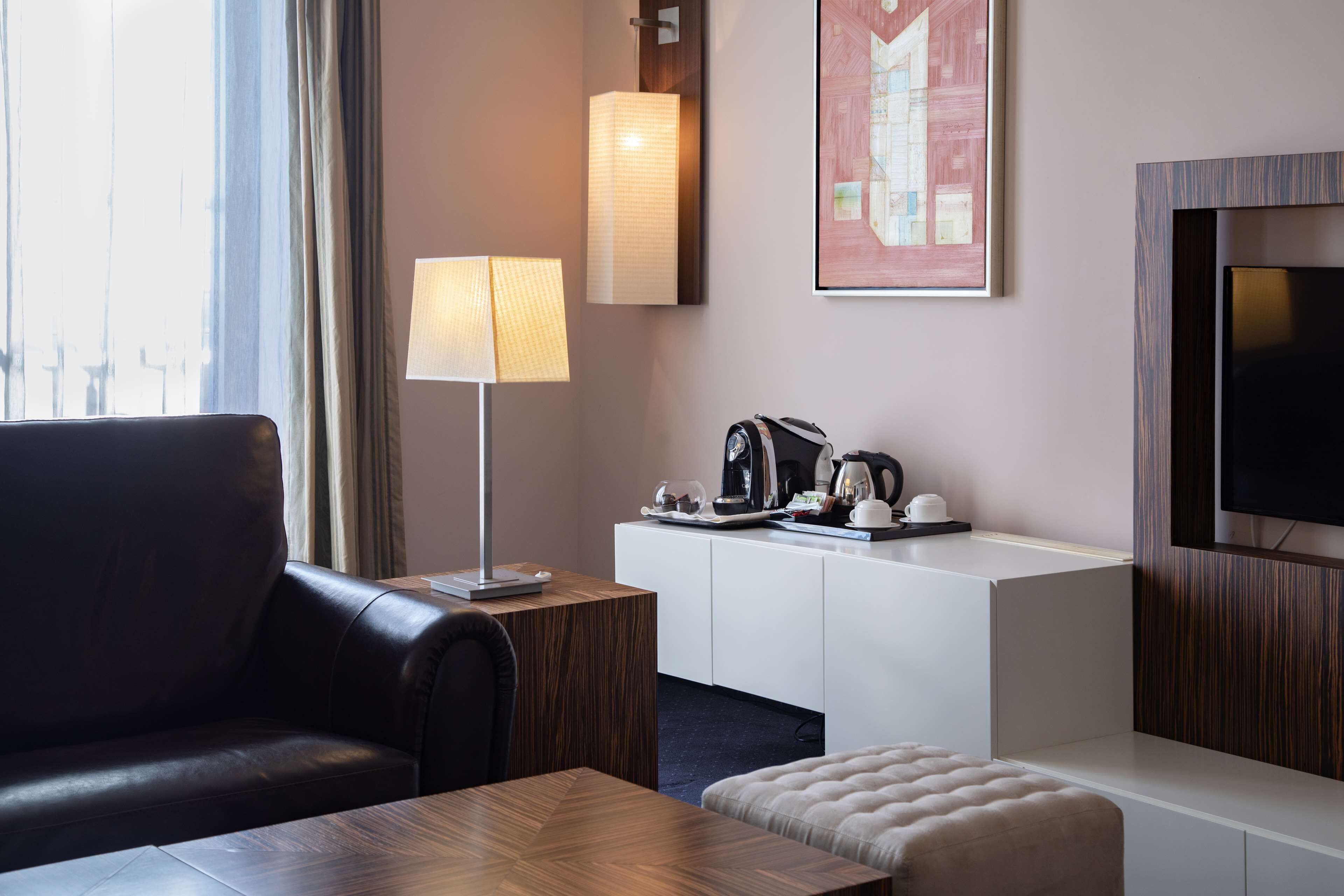 Une chambre d’hôtel moderne avec un canapé en cuir, une table basse en bois, un meuble blanc contenant une machine à café, une lampe et un téléviseur à écran plat.