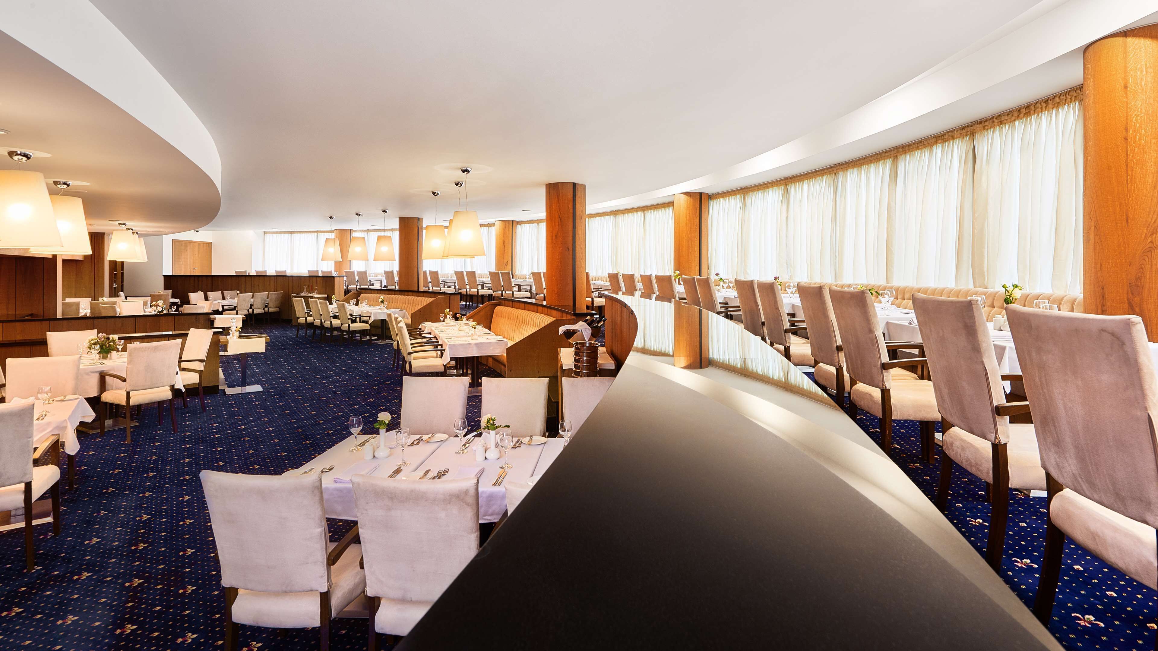 Een hotel eetruimte met meerdere tafels en stoelen, een gebogen zithoek en een blauw tapijtvloer.