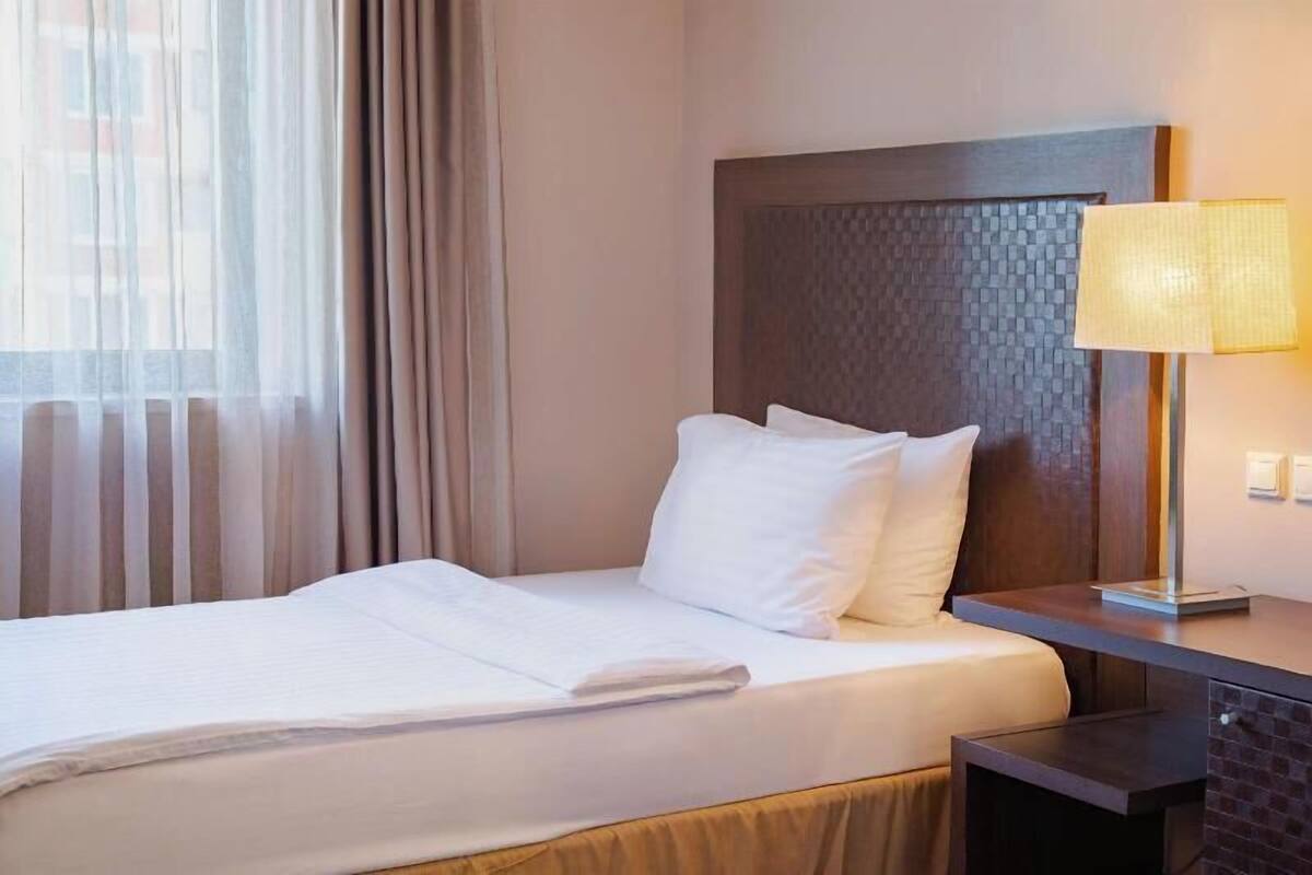 A hotel room with a bed, a nightstand, a lamp, and a view of the outside through sheer curtains.