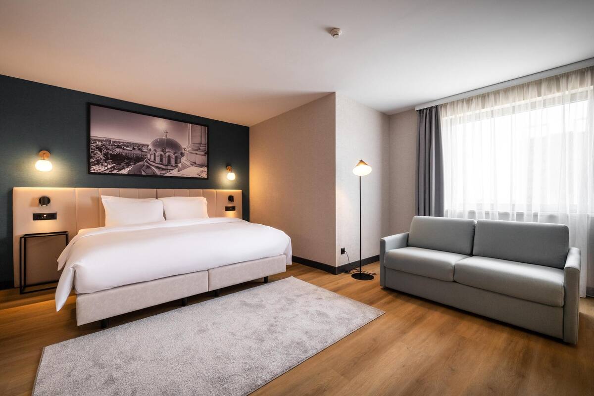 A modern hotel room with a large bed, a grey sofa, a framed cityscape picture on the wall, and a floor lamp.