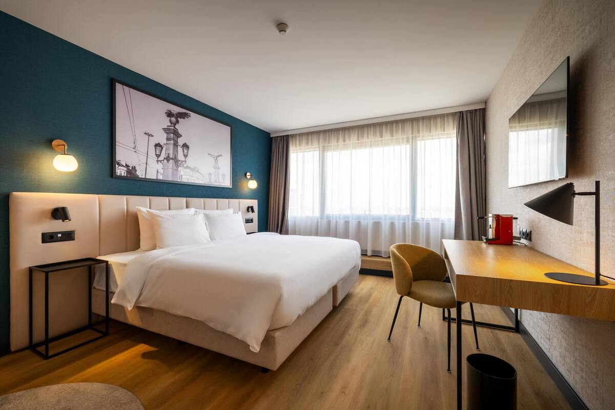 A modern hotel room with a large bed, a desk, and a chair. There is a painting on the wall, a window with curtains, and a ceiling light.