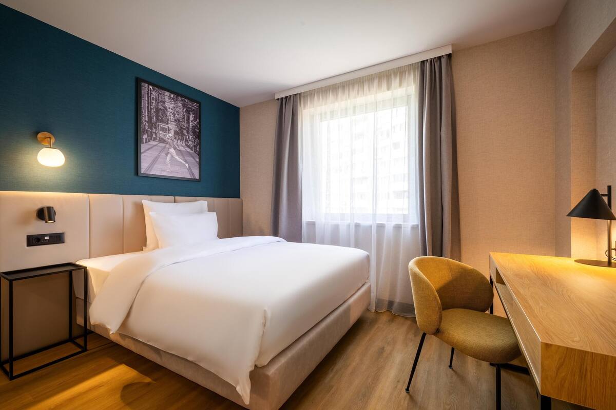 A hotel room with a bed, a desk, a chair, a nightstand, a wall-mounted light, and a window with curtains.