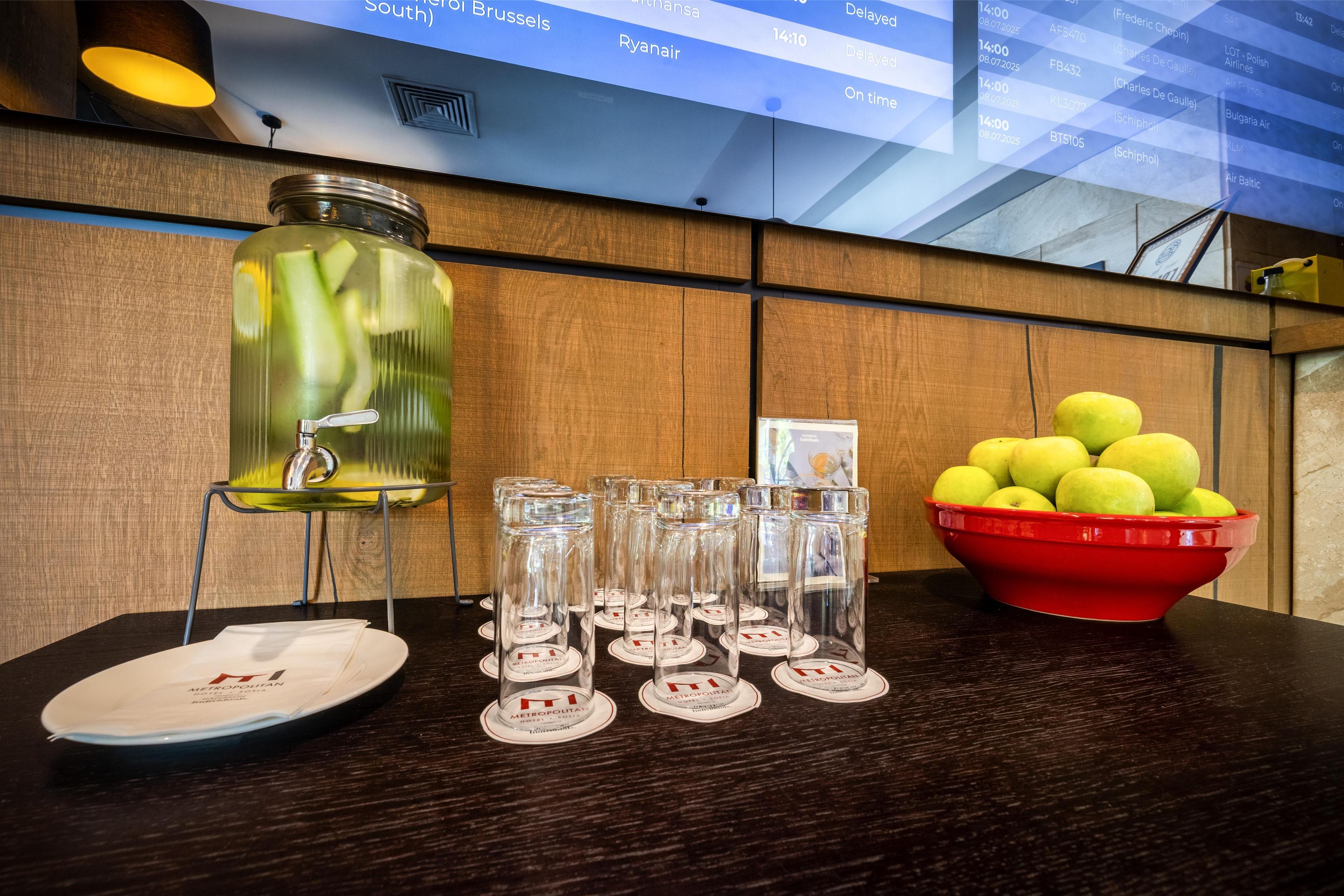 Une table avec un bocal en verre, plusieurs verres, un saladier de pommes et, au fond, un panneau affichant le menu.