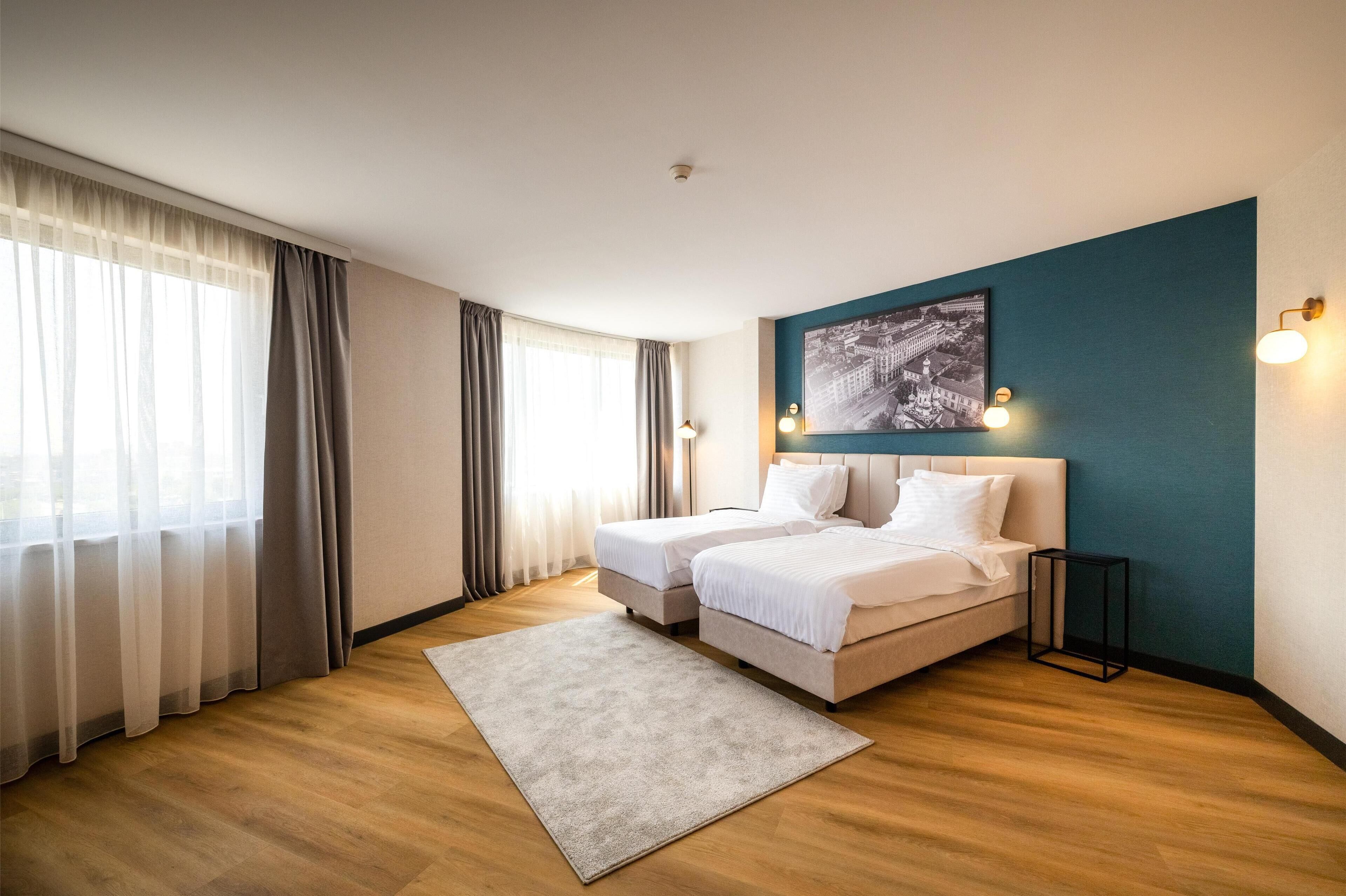 A hotel room with two beds, a large window with sheer curtains, a cityscape painting on the wall, and a small side table.