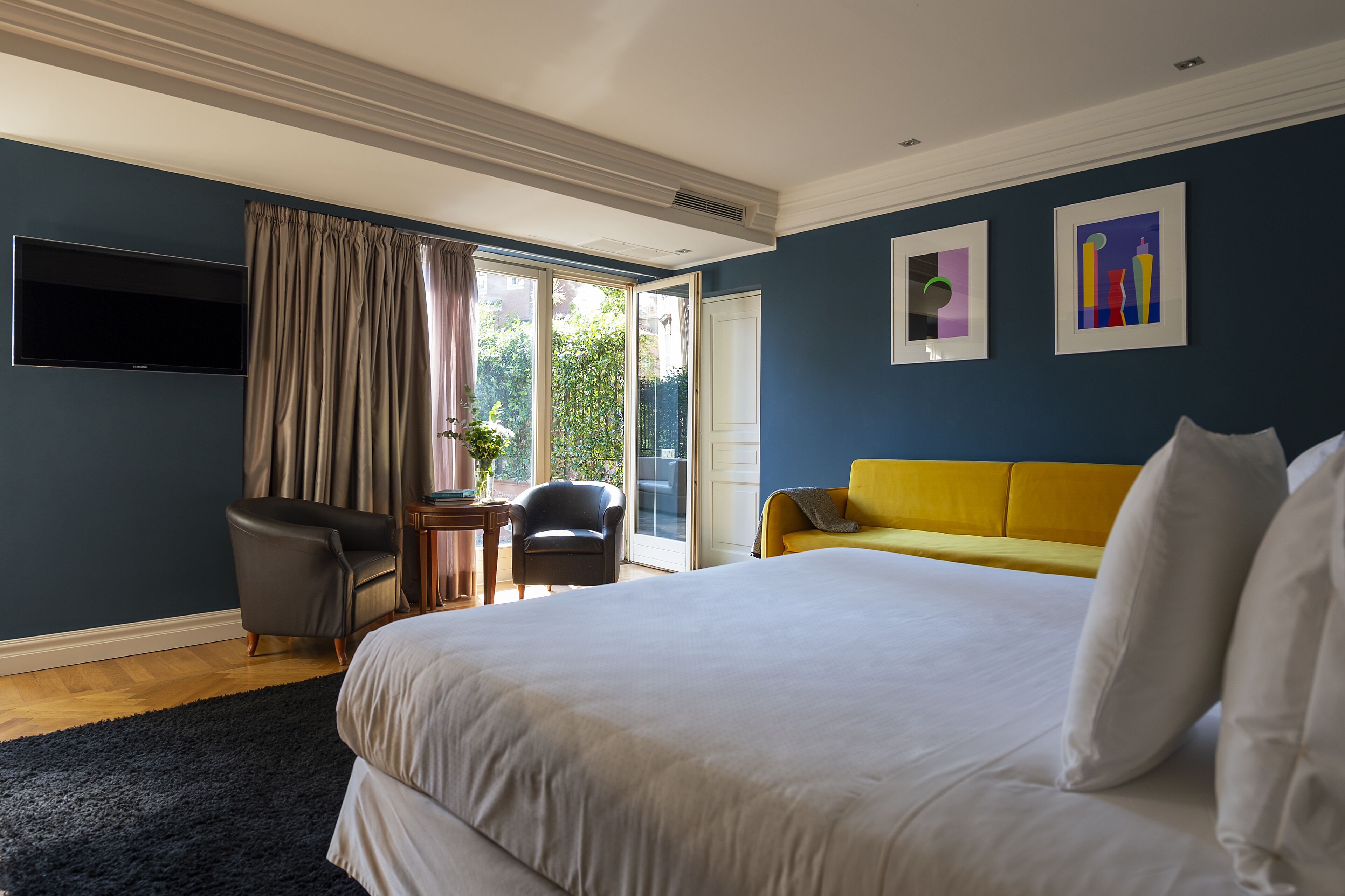 Una camera d'albergo con un letto grande, un divano giallo, una TV fissata alla parete e una porta finestra scorrevole che dà su un balcone.