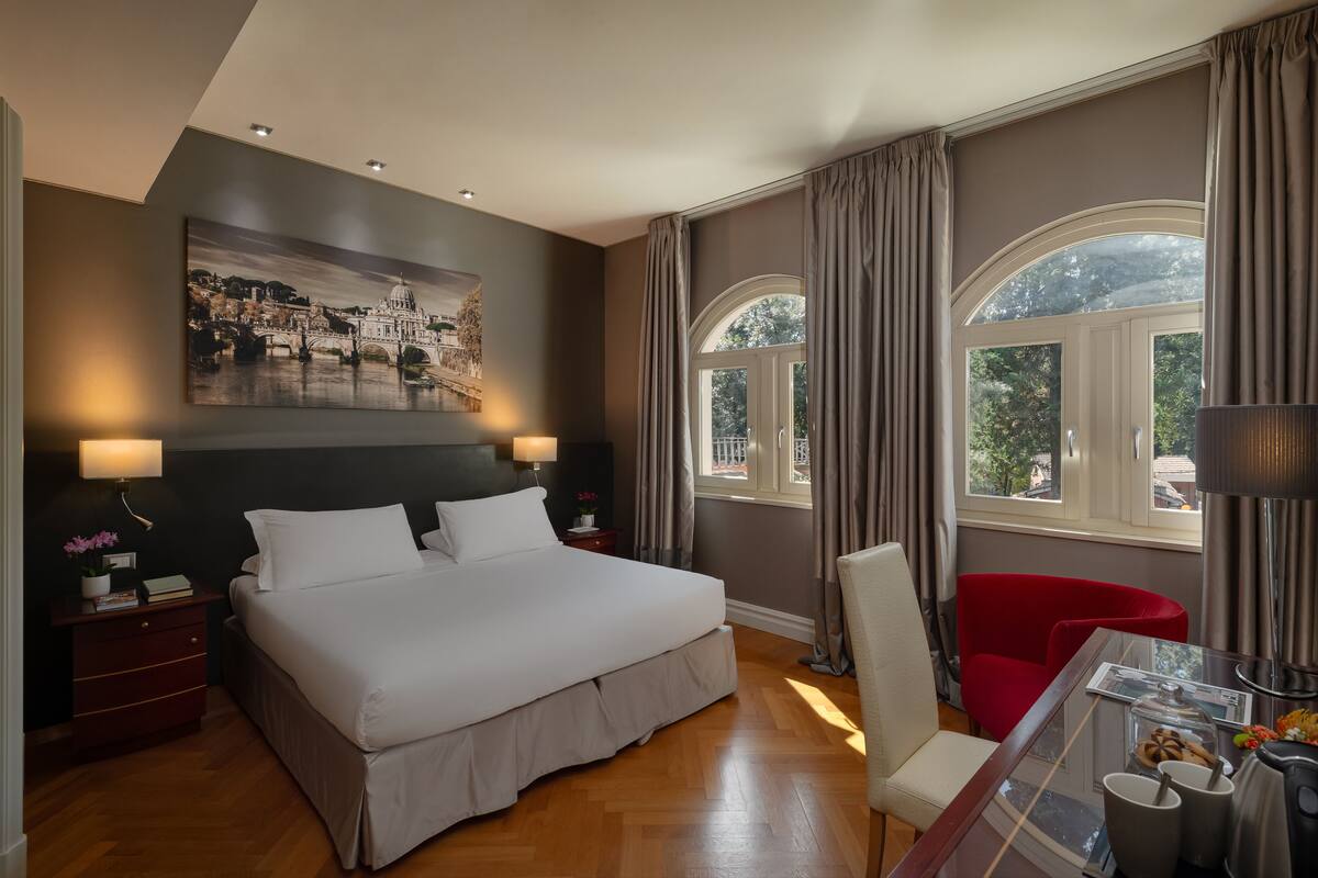 Habitación de hotel con una cama grande, un escritorio con silla, una ventana con cortinas y un cuadro en la pared.