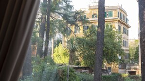 Une vue à travers la fenêtre d’un bâtiment donnant sur un jardin avec un chemin, de grands arbres et un autre bâtiment à la façade jaune.