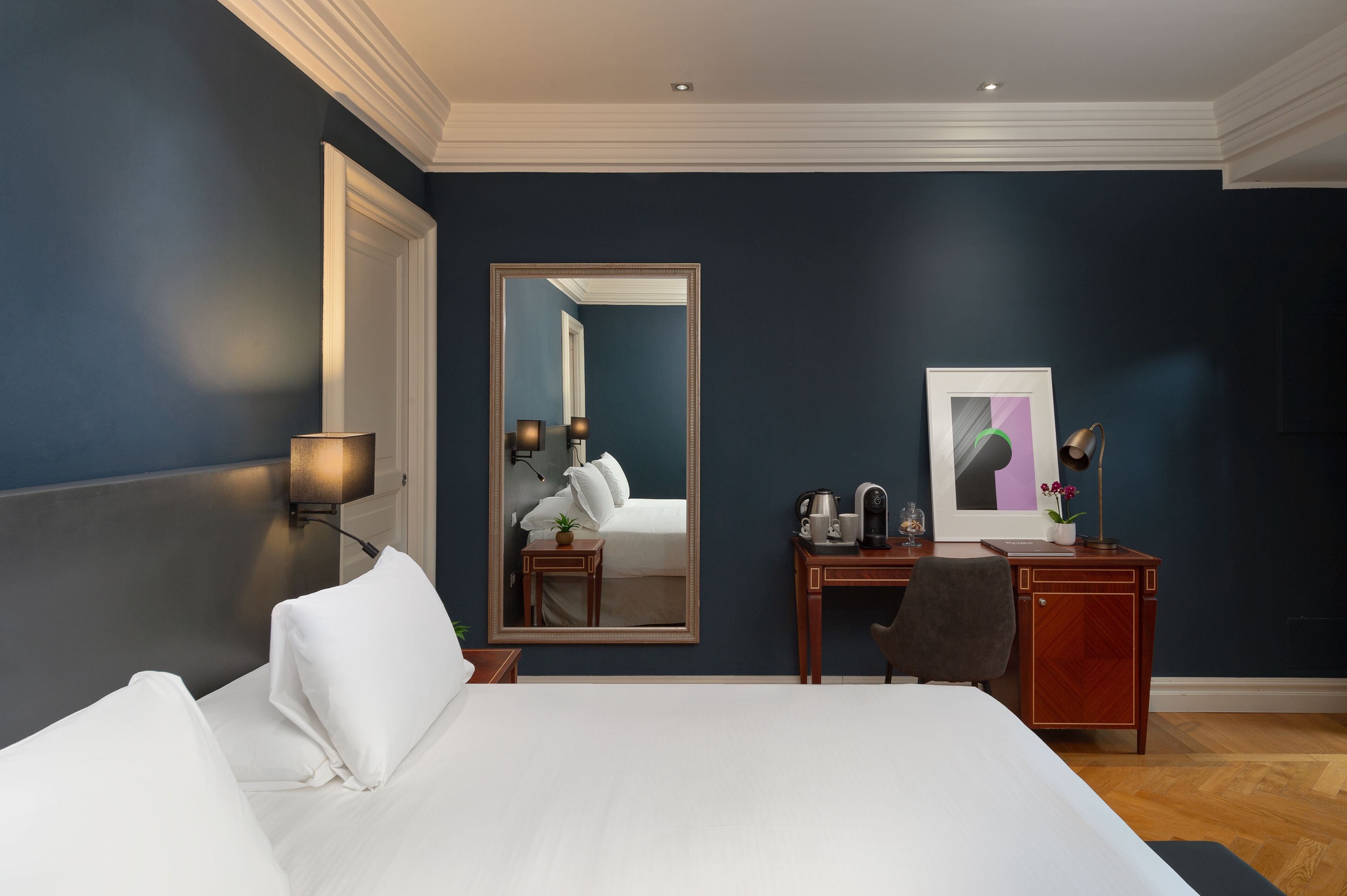 A hotel room with a large bed, a desk, a chair, a desk lamp, a mirror, and a framed picture on the wall.