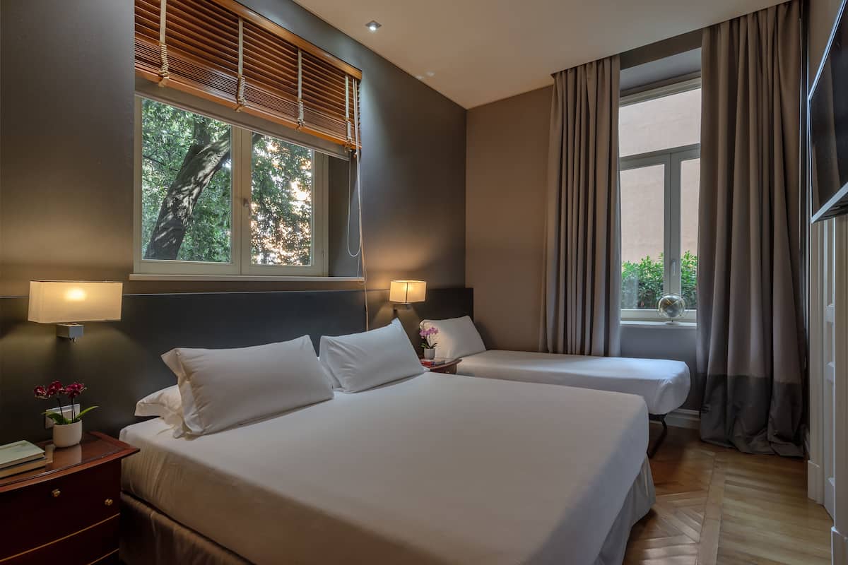 Habitación de hotel con dos camas, una mesita de noche con lámpara, una ventana con persianas y vistas al exterior con vegetación.