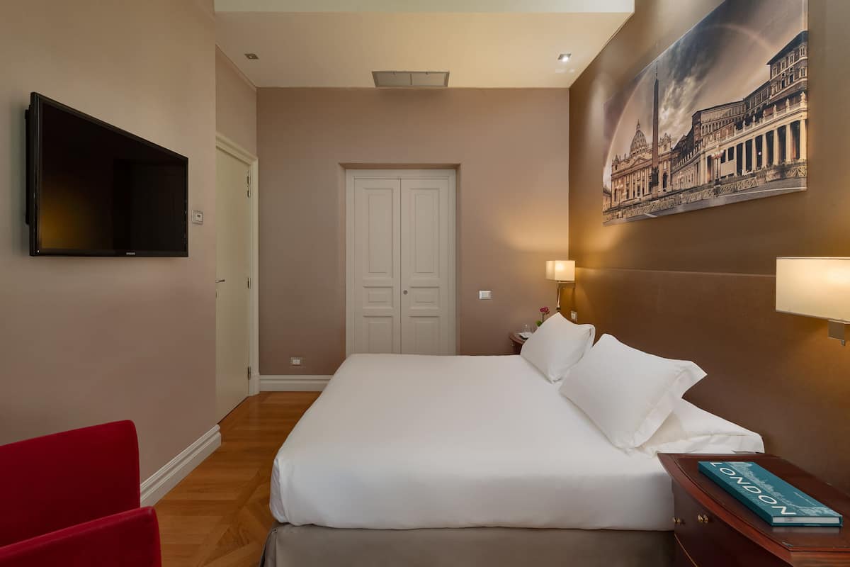 Habitación de hotel con cama, una silla roja, televisión de pantalla plana, una puerta y un cuadro de un edificio.