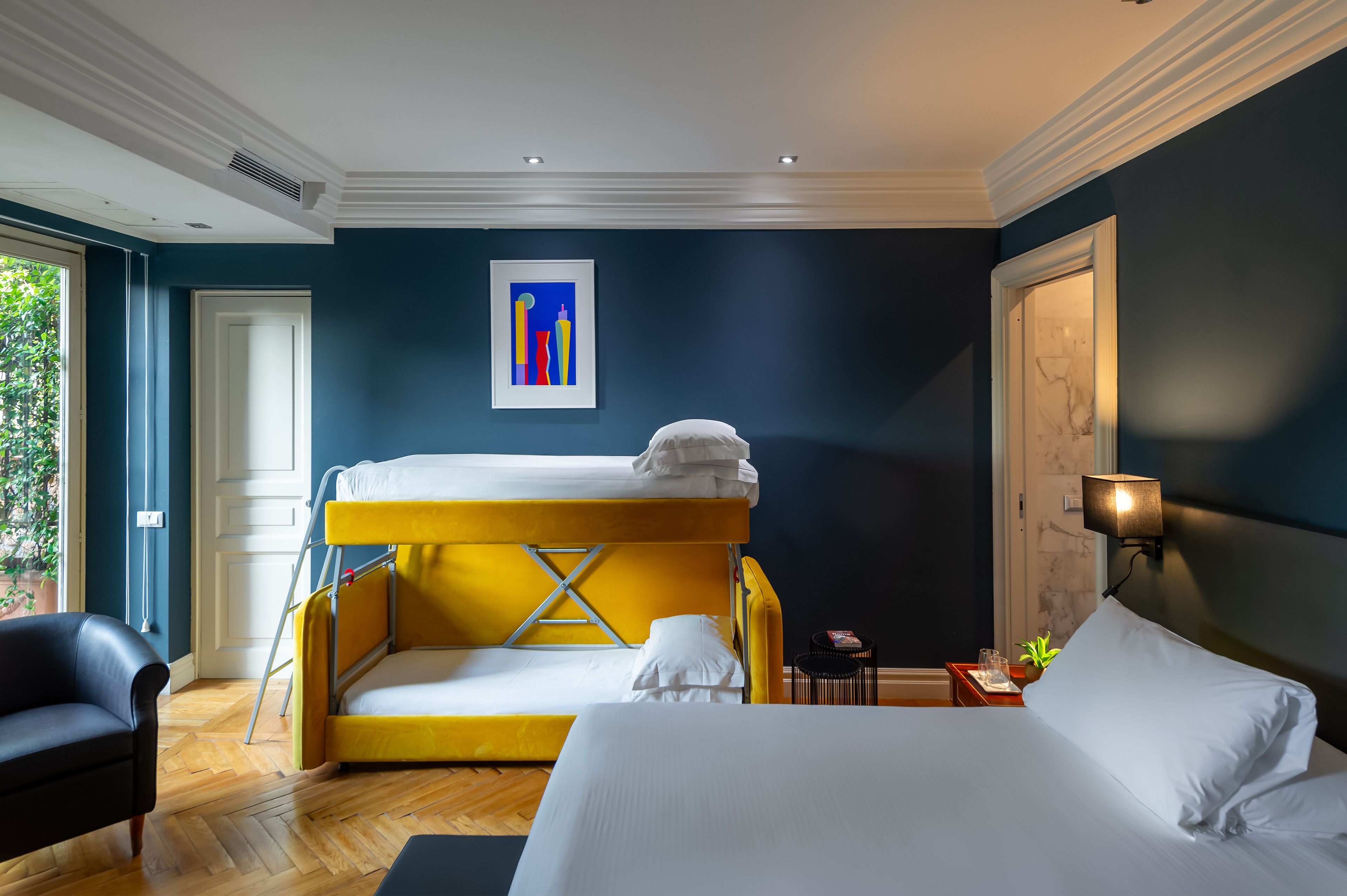 A hotel room with a bunk bed, a single bed, a chair, a small table with a lamp, and a framed artwork on the wall.