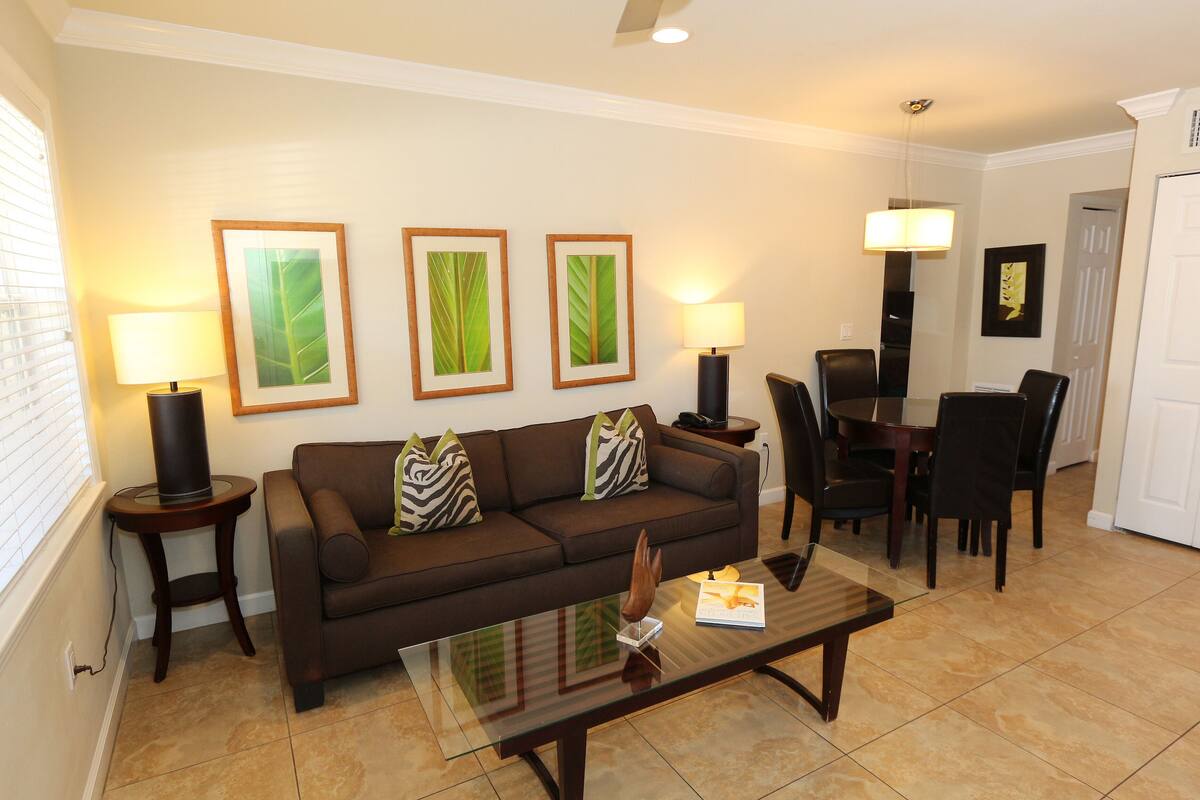A living room with a brown sofa, two side tables with lamps, a glass coffee table, and framed leaf artwork on the wall.