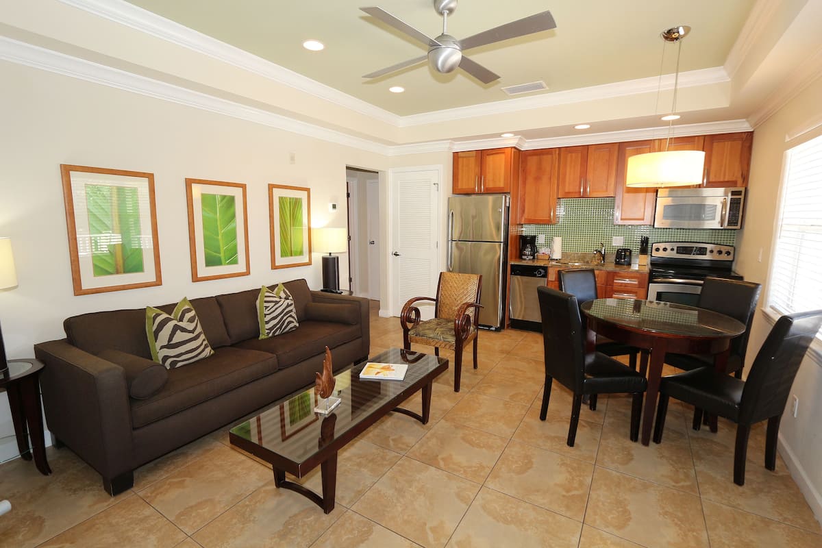 A living room with a sofa, coffee table, and chairs, a dining area with a table and chairs, and a kitchen with appliances and wooden cabinets.