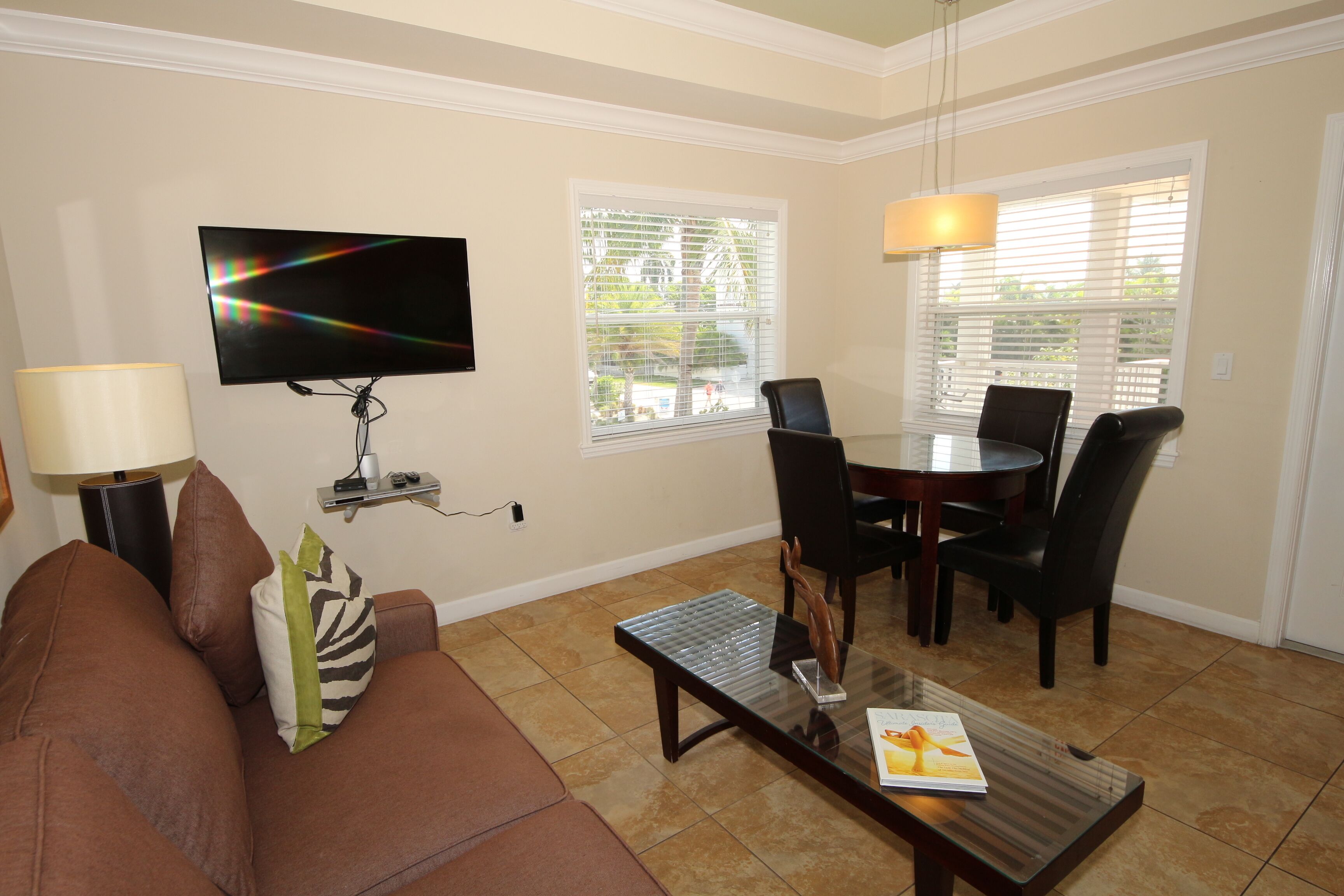 Una sala de estar con un sofá marrón, una mesa de centro con una revista, un televisor montado en la pared y una zona de comedor con mesa y sillas.