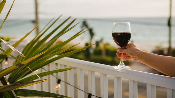 A hand holding a glass of red wine on a balcony with a view of the ocean. - The Beach Club at Anna Maria by RVA (Bradenton Beach)