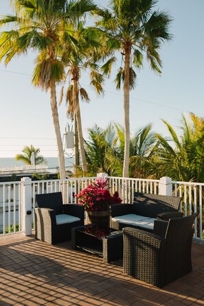 An outdoor patio with wicker furniture, a flower pot, and a view of the sea. - The Beach Club at Anna Maria by RVA (Bradenton Beach)