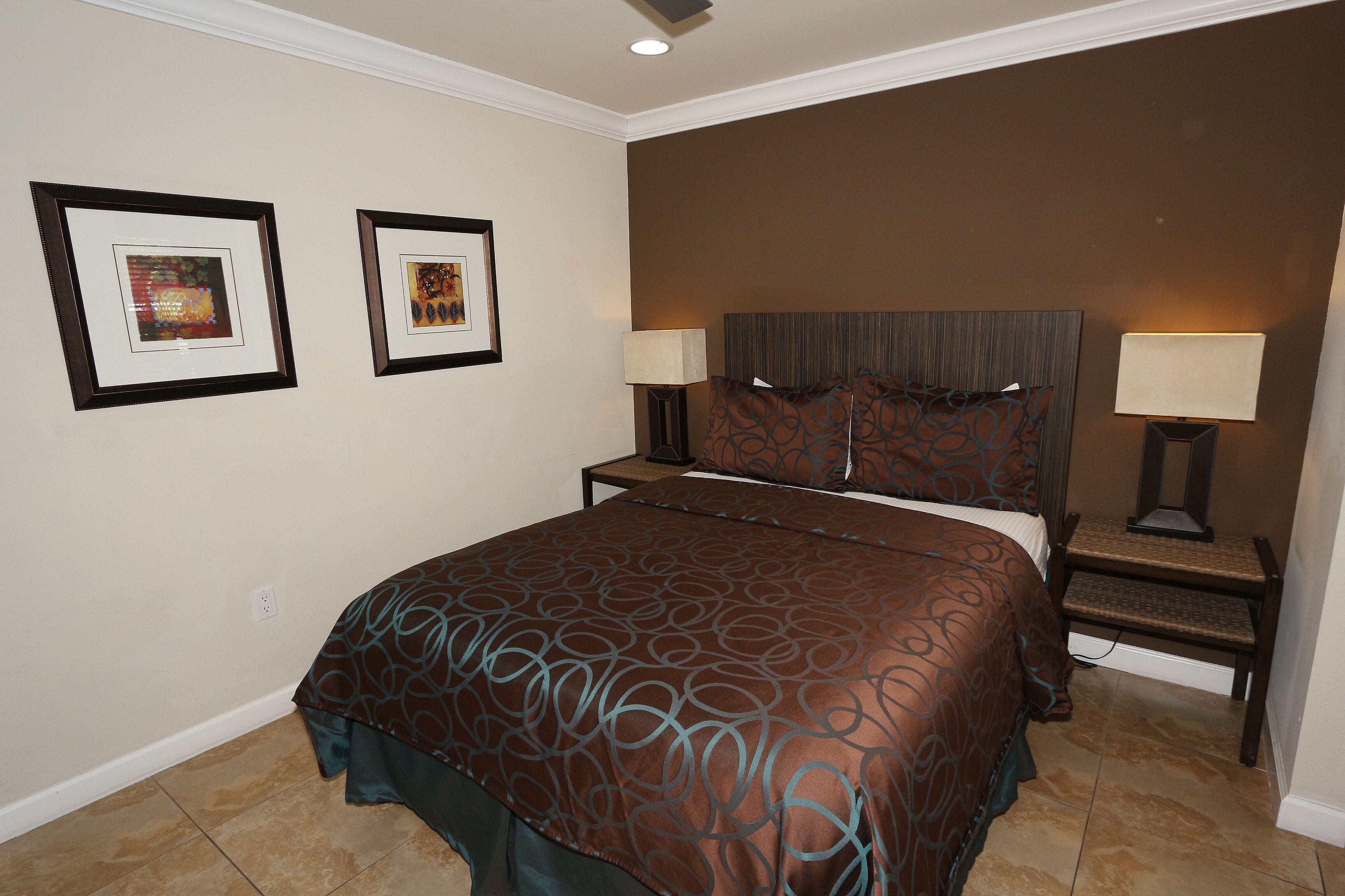 A hotel room with a large bed, two framed artworks on the wall, a nightstand with a lamp, and a headboard with a pillow.