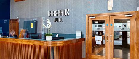 Una mesa de recepción con un letrero que indica Hotel Glashaus, una puerta de madera que dice "Osten Bar" y una planta en maceta.