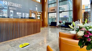 A hotel reception area with a wooden reception desk, seating area with orange chairs, and large windows offering views of the city.