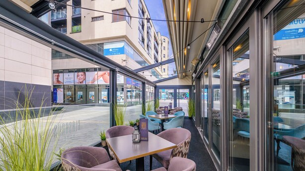 An outdoor seating area with cushioned chairs and tables, surrounded by glass walls and potted plants.
