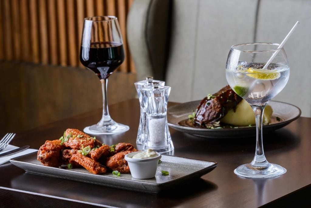 A dining table with a plate of chicken wings, a glass of red wine, a glass of water with a lemon slice, and a salt and pepper shaker.