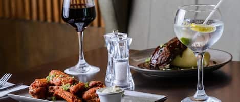Una mesa de comedor con un plato de alitas de pollo, una copa de vino tinto, un vaso de agua con una rodaja de limón y un salero y pimentero.