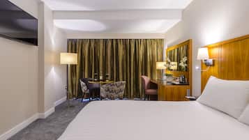 A hotel room with a large bed, a desk with a chair, a flat-screen TV mounted on the wall, and a large window with curtains.