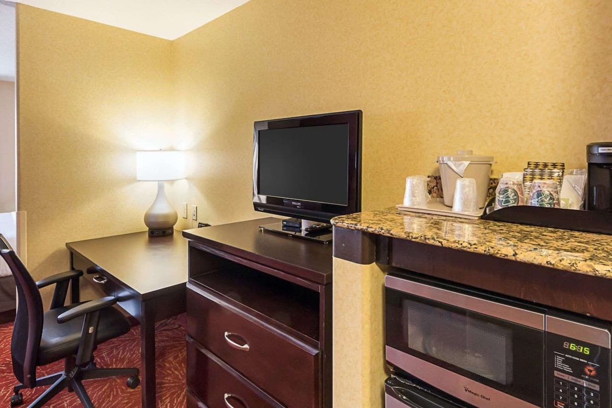 A hotel room with a desk, chair, television, microwave, coffee maker, and a small kitchenette area.