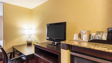 A hotel room with a desk, chair, television, microwave, coffee maker, and a small kitchenette area.