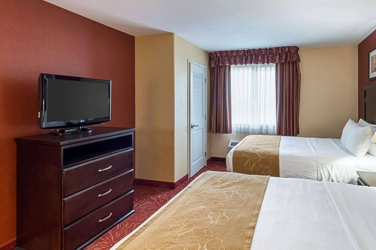 A hotel room with a flat-screen TV on a wooden dresser, two beds with white linens, a large window with curtains, and a red carpet.