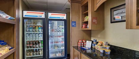 Una kitchenette pequeña con dos refrigeradores llenos de bebidas y aperitivos, una encimera con diversos alimentos, y un estante de pared con un cartel de aperitivos.