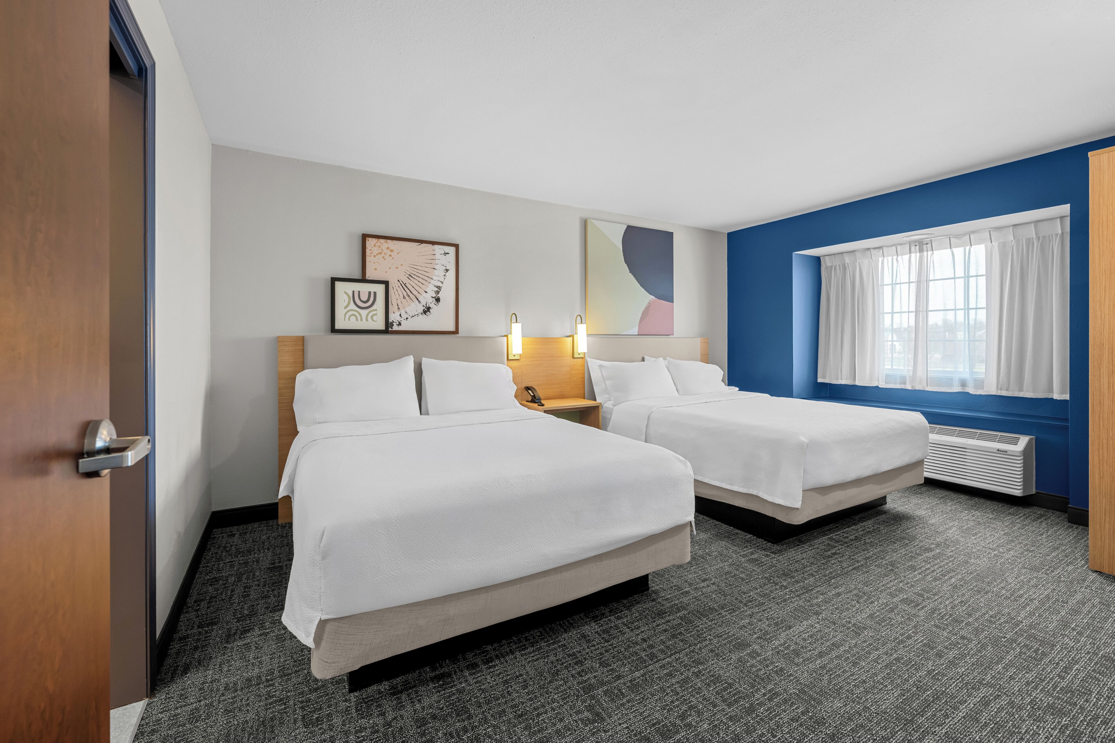 A hotel room with two beds, a wooden headboard, a wall-mounted lamp, a framed picture, and a window with curtains.