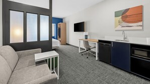 A modern hotel room with a sofa, desk, kitchenette, and a large window with blinds. - Spark by Hilton Stillwater (Stillwater)