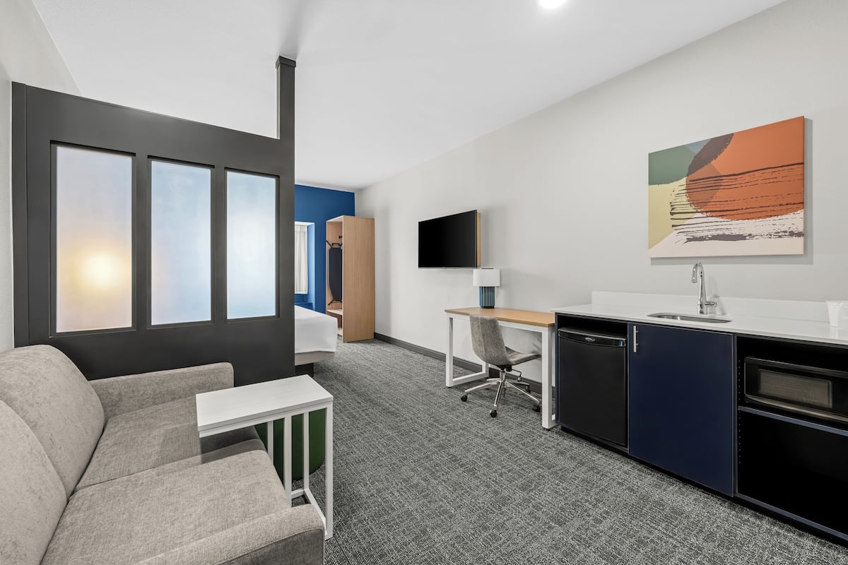 A modern hotel room with a sofa, desk, kitchenette, and a large window with blinds.