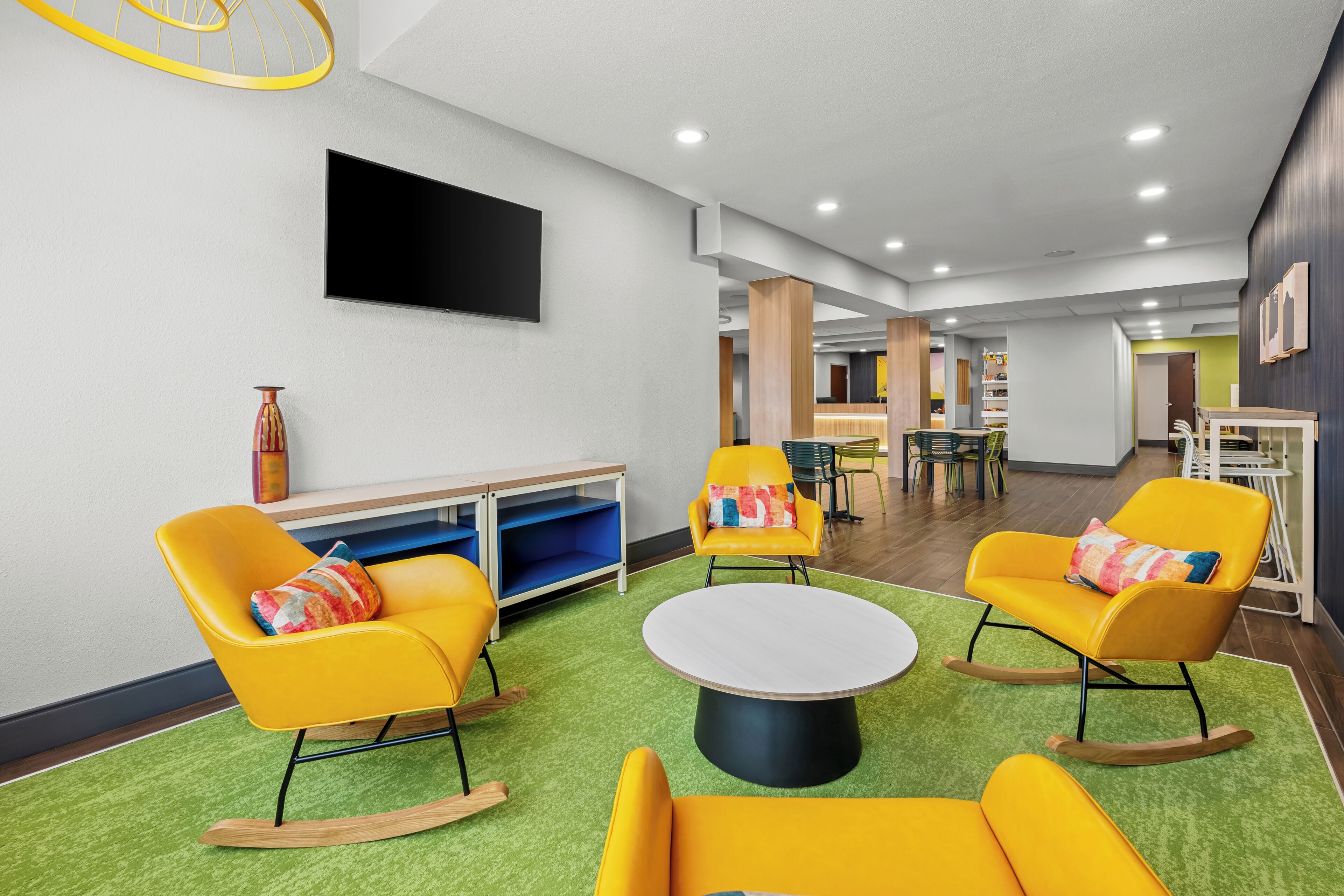 Un hall d’hôtel moderne avec des fauteuils à bascule jaunes, une table ronde et un téléviseur à écran plat fixé au mur.
