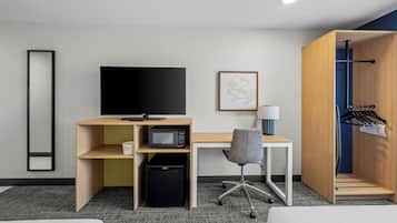 Habitación de hotel con escritorio, televisión, microondas, basurero, silla, perchero y cama.