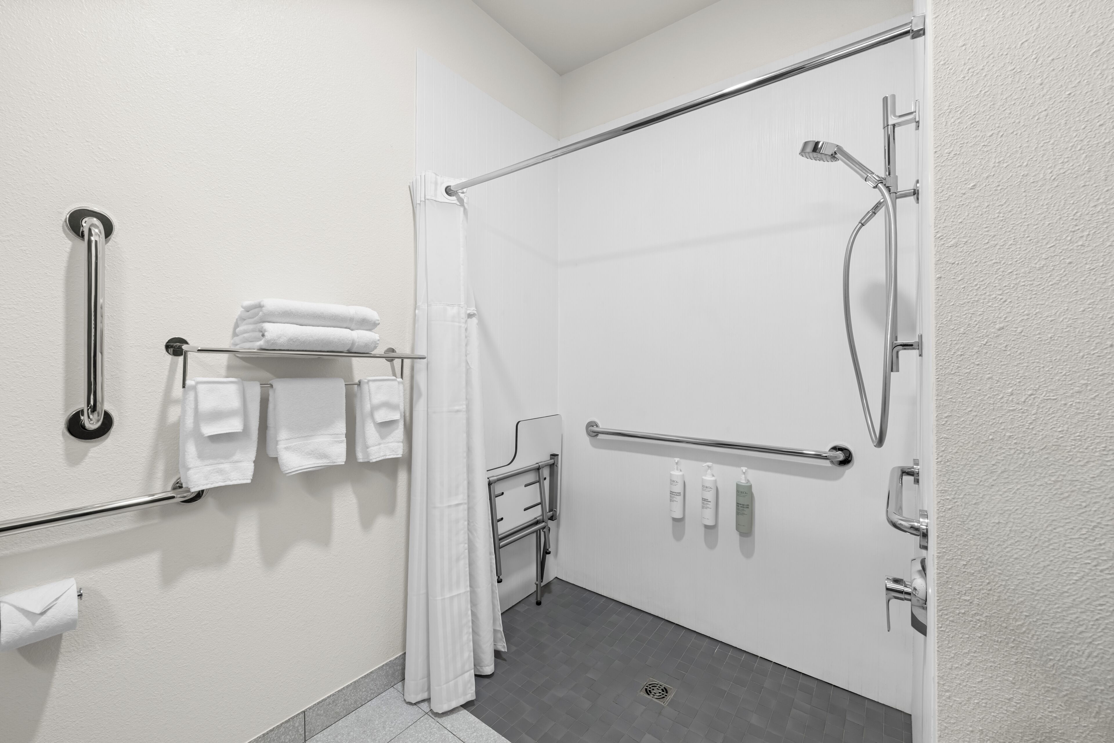 Un baño con ducha, barras de apoyo, toallero y dispensador de papel higiénico.