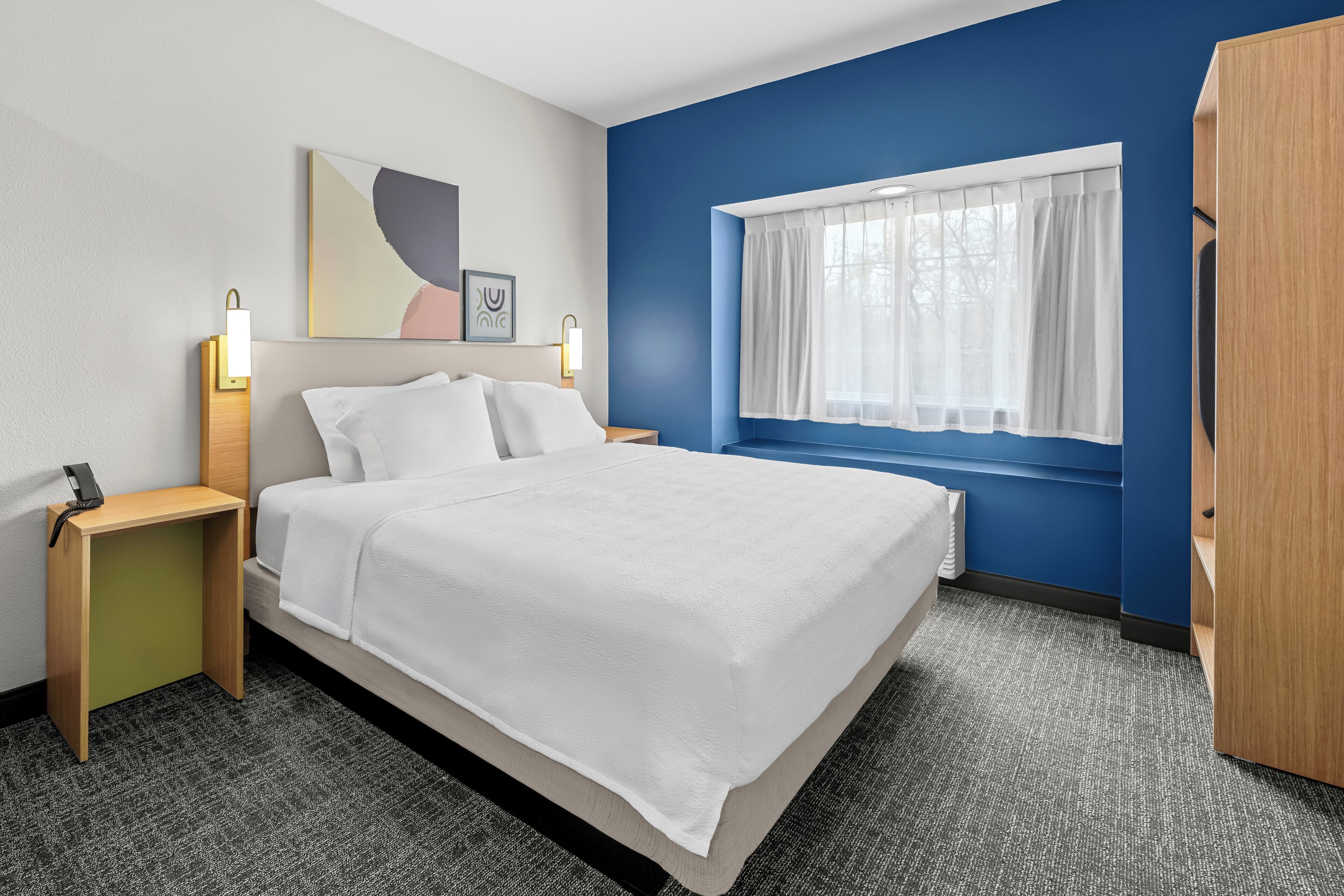 A hotel room with a bed, bedside tables, a window with curtains, a wall painting, and a wardrobe.