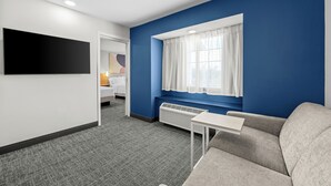 A hotel room with a flat-screen TV, a window with white curtains, a gray sofa, a small table, and a white bedside table. - Spark by Hilton Stillwater (Stillwater)