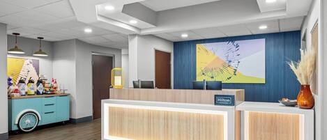 A hotel reception area with a wooden reception desk, a blue wall with a mural, a green carpet, and a blue counter with amenities.