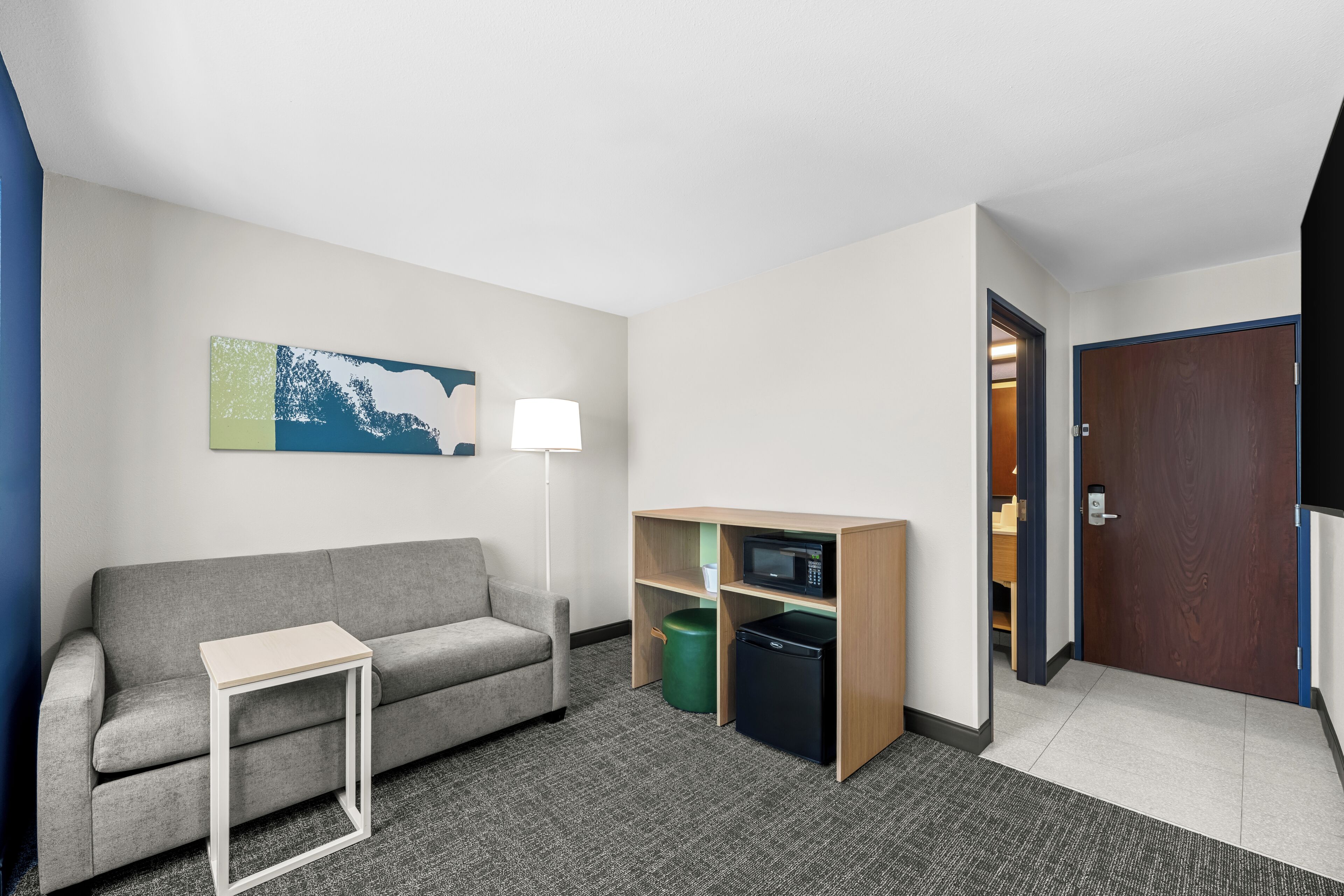 Ein Hotelzimmer mit grauem Sofa, einem kleinen weißen Tisch, einer Mikrowelle und einem kompakten Kühlschrank.