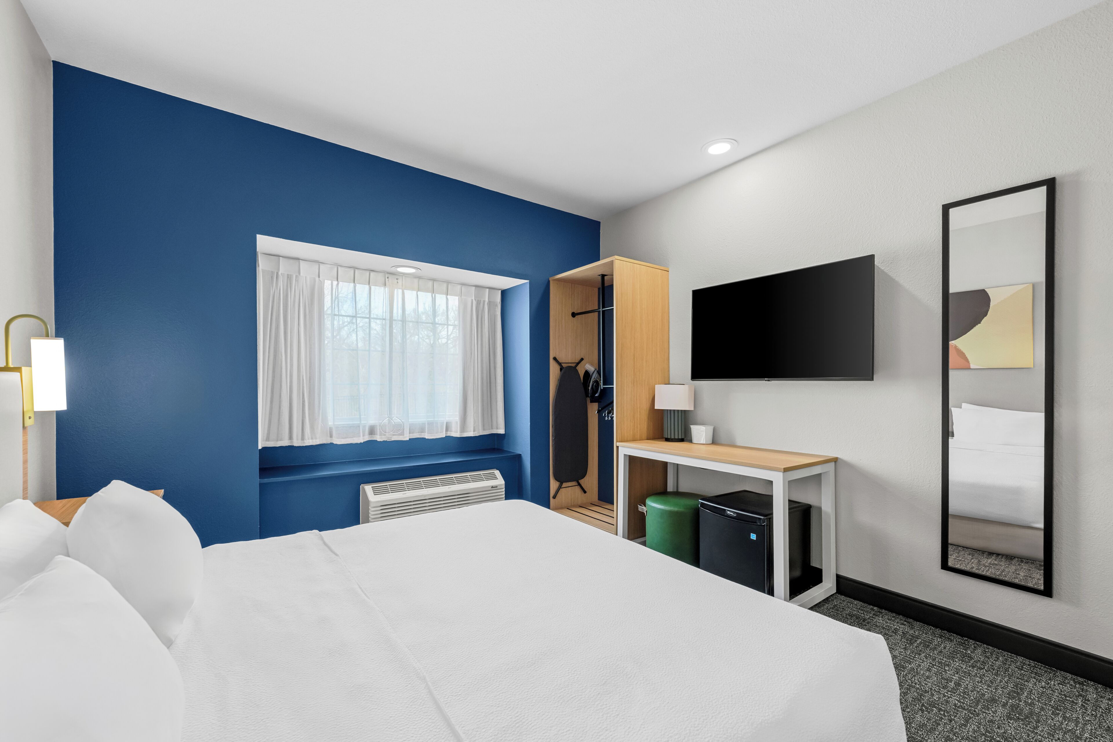A hotel room with a large bed, a desk, a mirror, a TV, and a window with curtains.