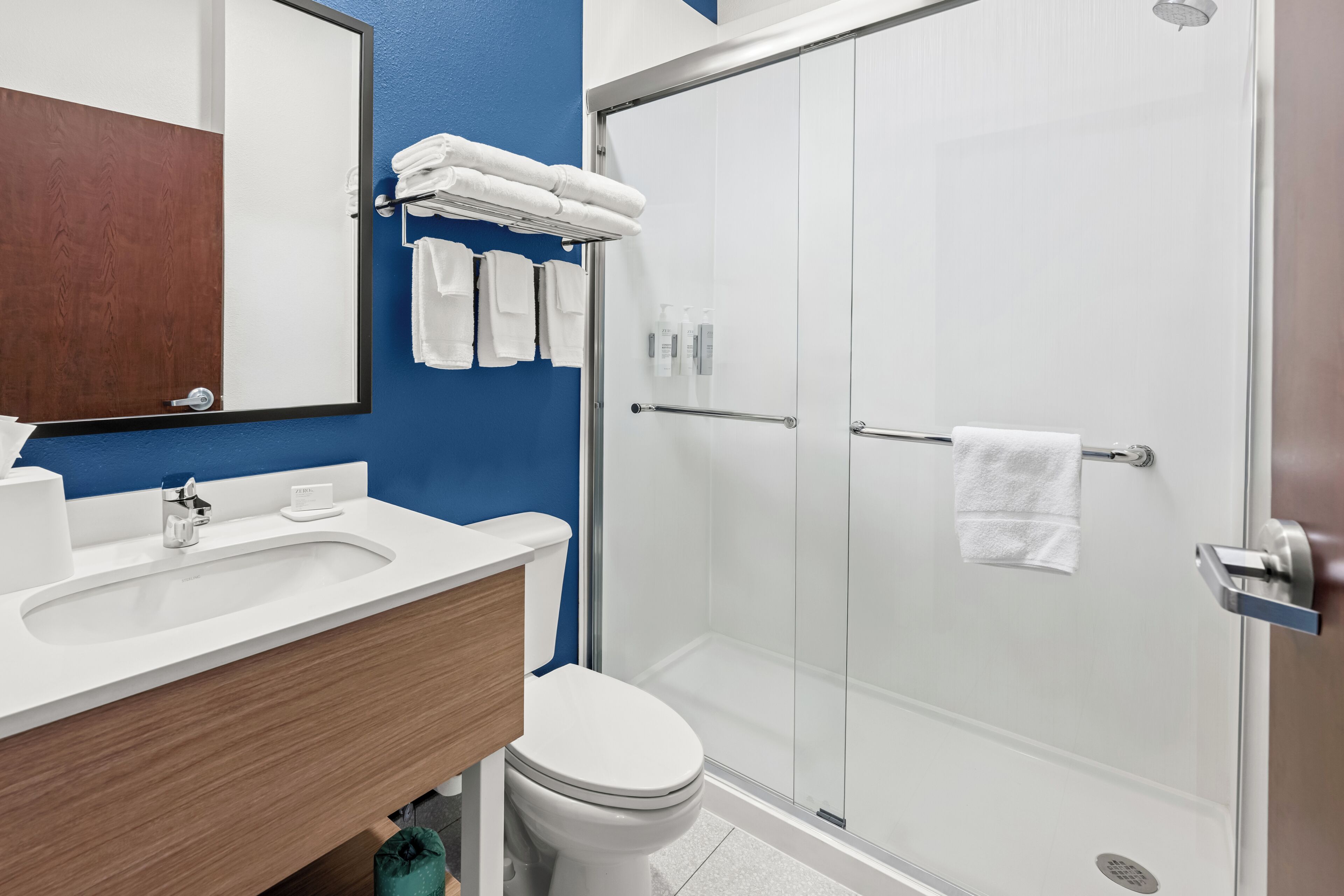 Een badkamer met een wit wastafelmeubel, een toilet, een douchecabine met glazen wanden en een blauwe wand.